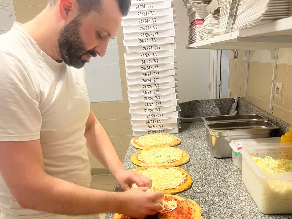Tugdual Boutillier parsemant de fromage râpé des pizzas à la sauce tomate alignées sur le plan de travail, devant une pile de boîtes « 24/24 - 7/7 »